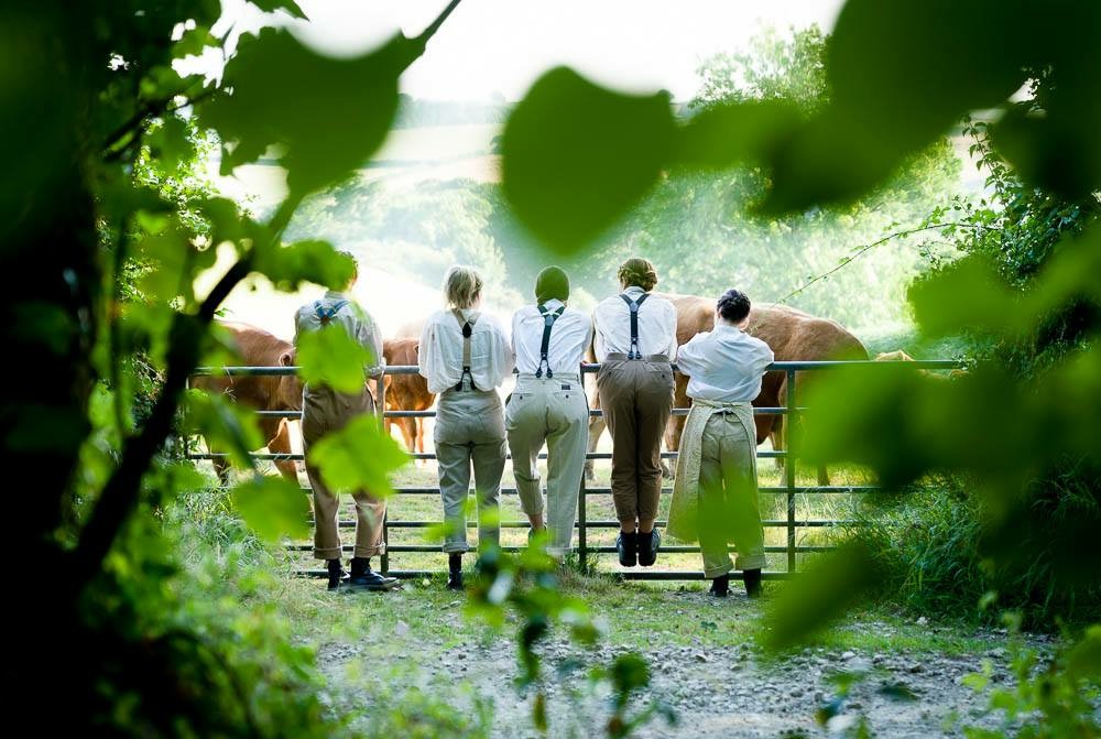 Five performers in period farm clothing lean on a gate watching cattle in a sunlit field, framed by green leaves