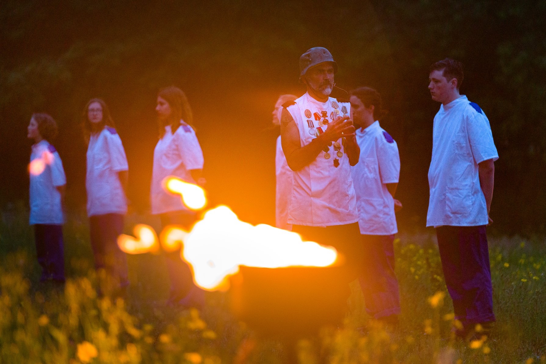 A performer in a medal-covered vest and helmet stands before a plume of fire, flanked by performers in white