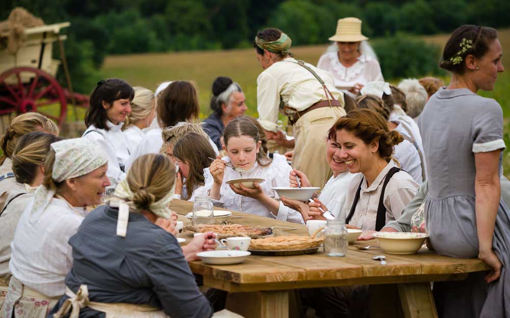 A large group in period dress shares food at a long wooden table in a field, an old cart visible behind them