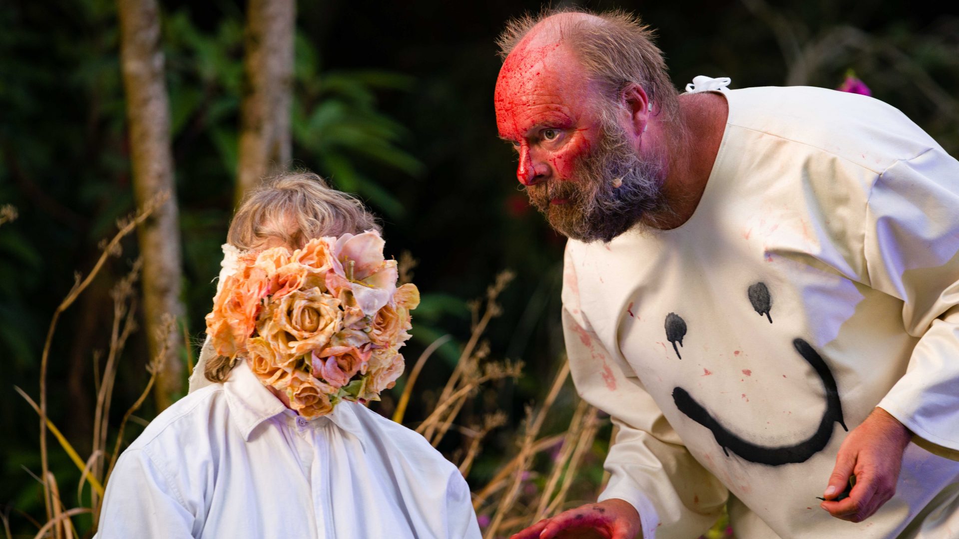 A performer with a blood-red painted face leans toward a performer whose face is covered by a mass of faded roses