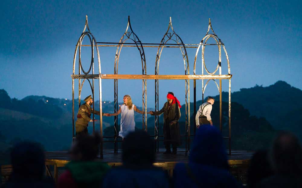 Performers stand within a tall metal frame structure on a hilltop stage at dusk, silhouetted audience heads in the foreground