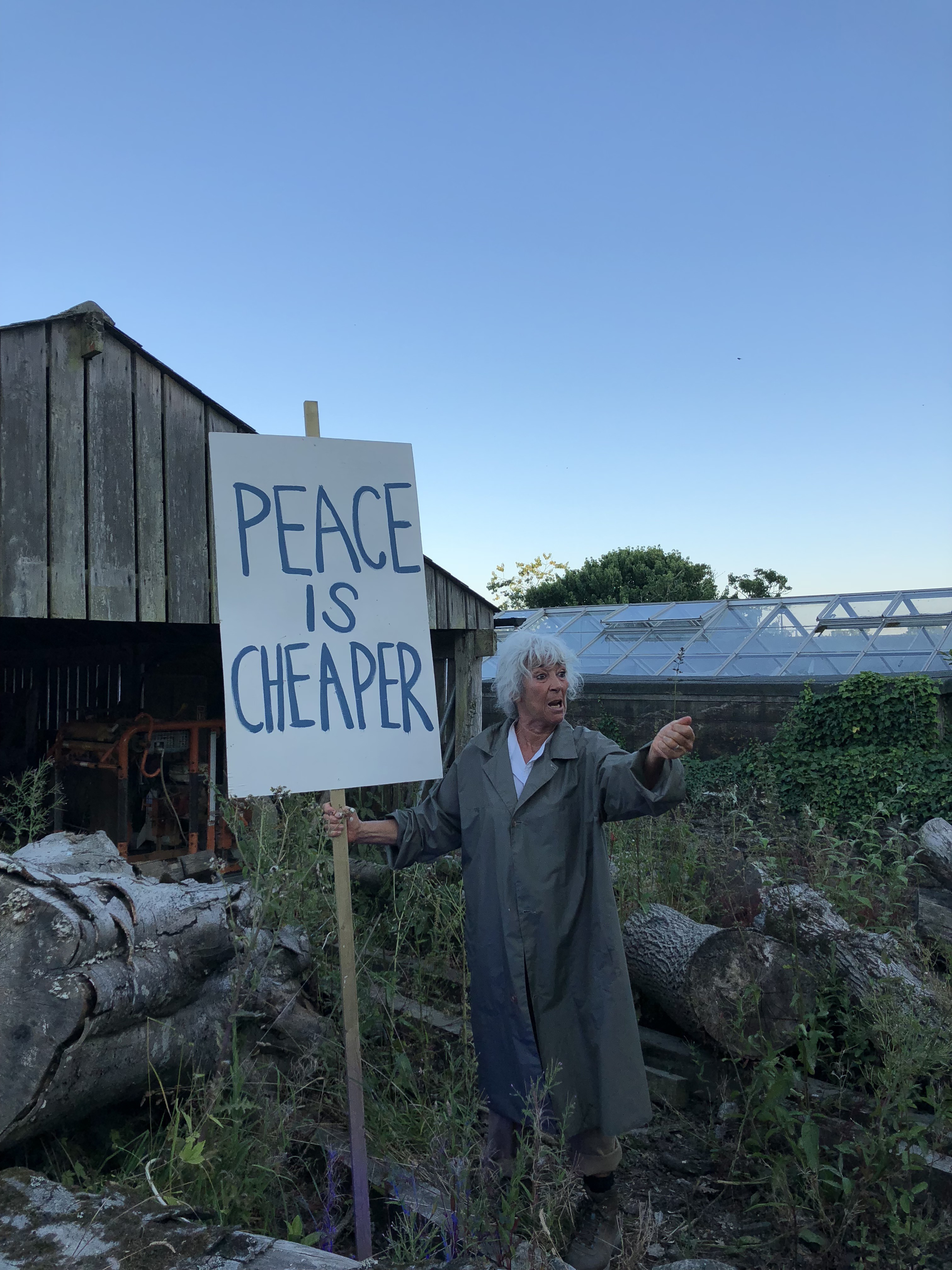 A performer in a raincoat holds a placard reading Peace Is Cheaper in front of a barn and greenhouse