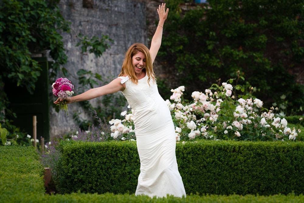 A performer in a white dress raises a bouquet in celebration in a walled garden of roses