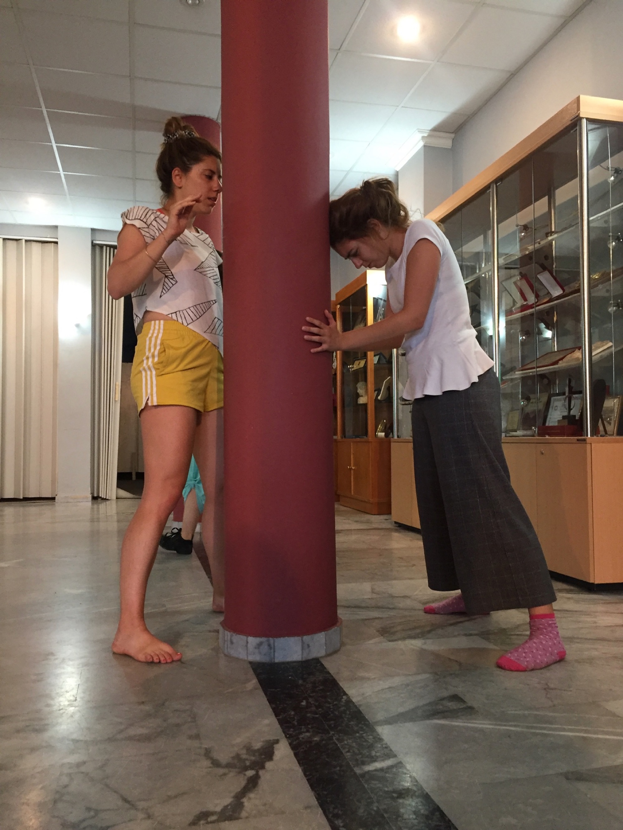 Two performers face each other across a red column in a marble-floored foyer lined with glass display cases