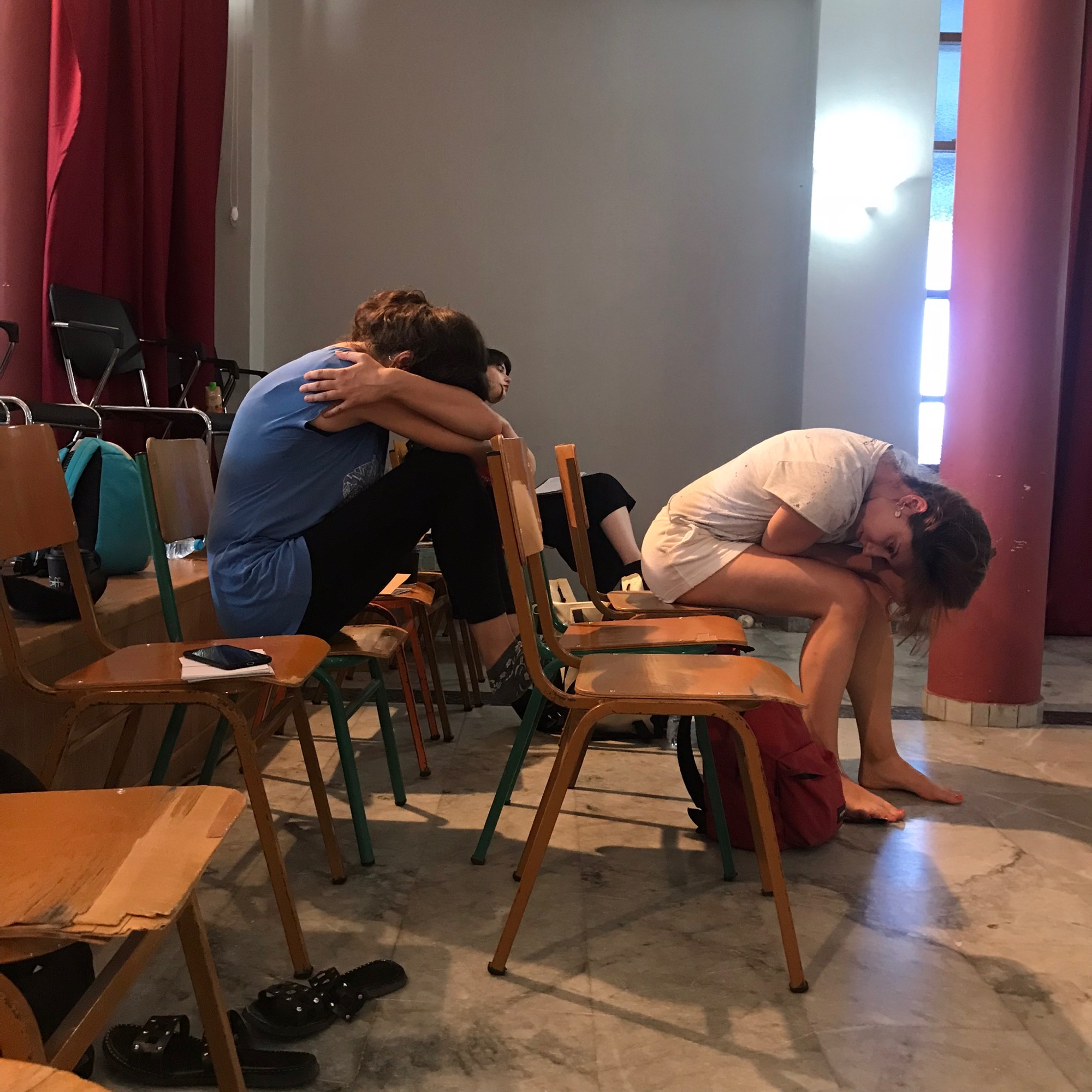 Two performers rest draped over rows of stacked classroom chairs in a rehearsal room strung with red curtains