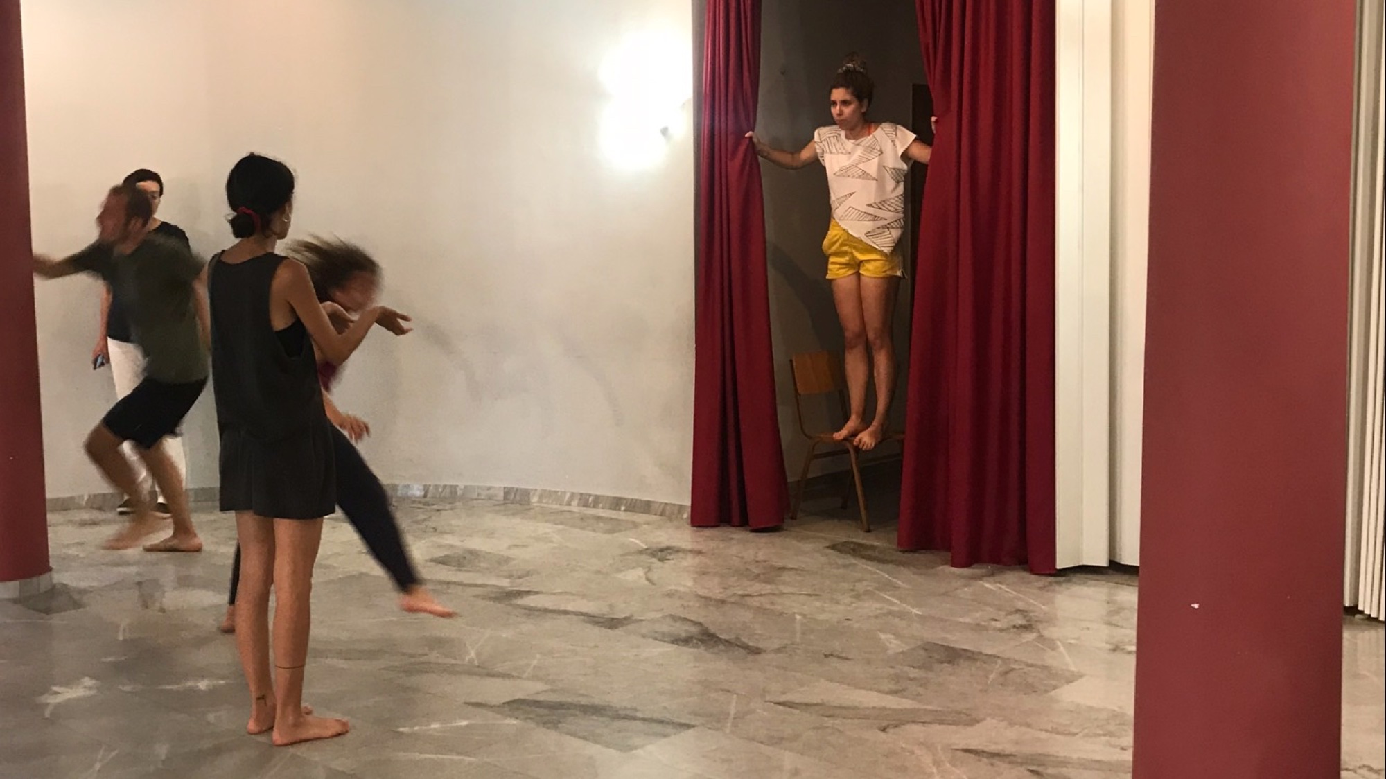A performer stands on a chair pulling back a heavy red curtain in a doorway as others move across the marble floor in the foreground
