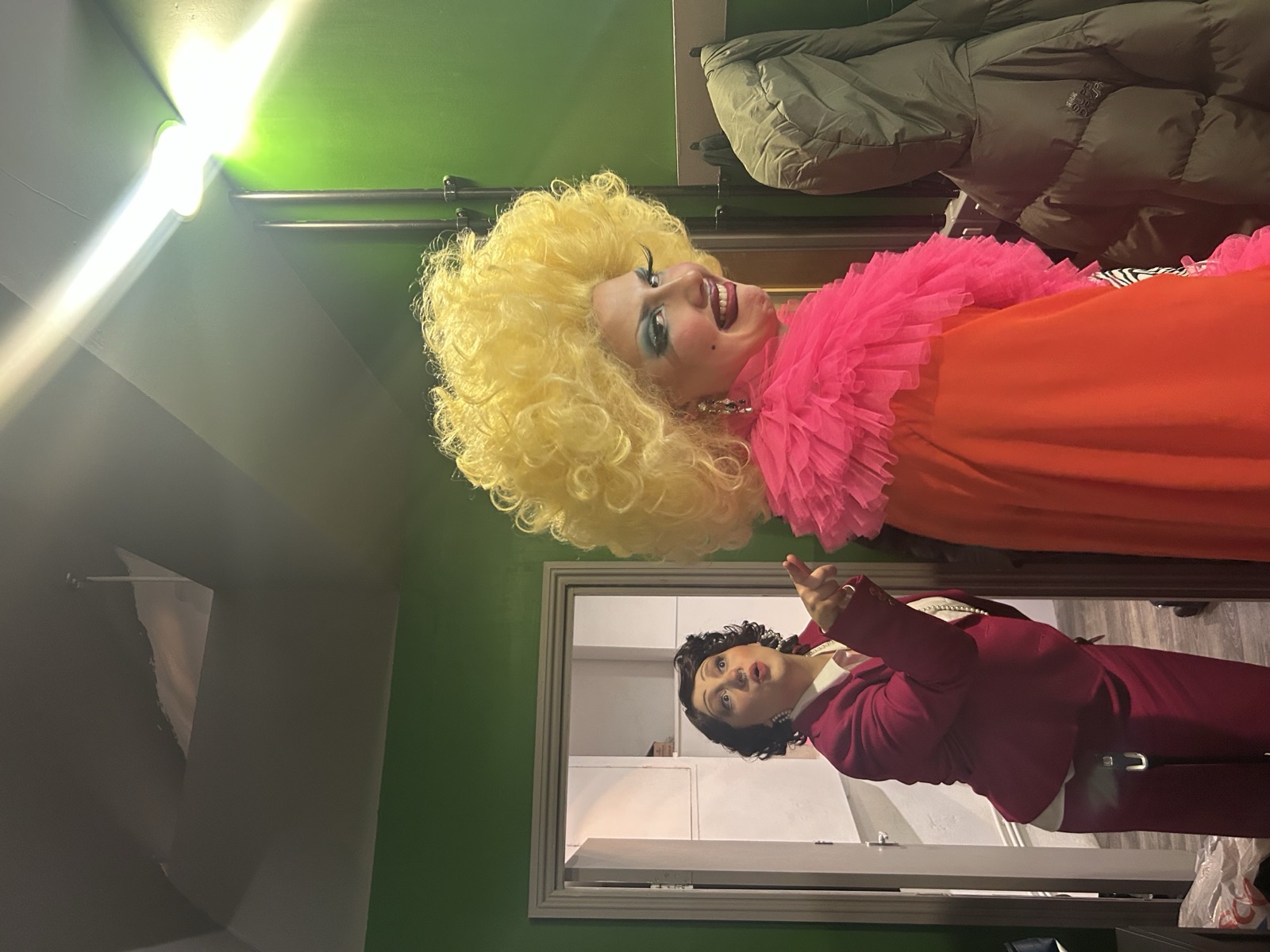 Cis-Celeste backstage in a big yellow curly wig, pink ruffled collar, and orange cape, with a co-star in a maroon suit in the mirror