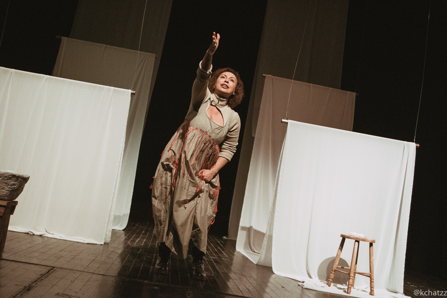 A performer in a beige dress trailed with red thread reaches an arm upward toward the light, framed by hanging white curtain panels