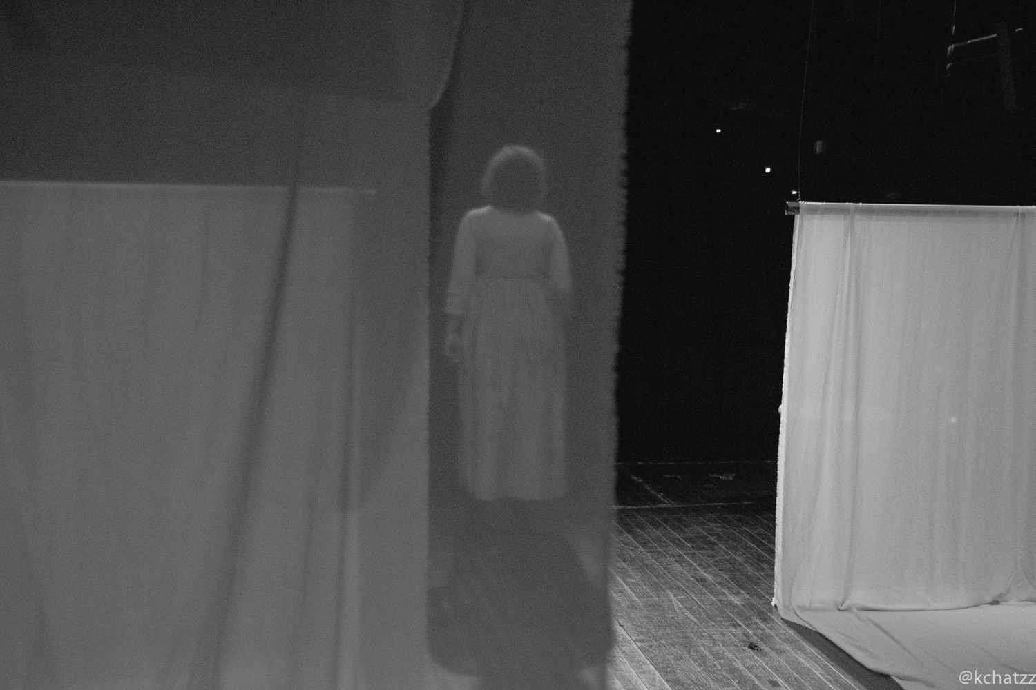 A blurred silhouette of a woman in a white dress and curly hair glimpsed through a gap in white fabric panels