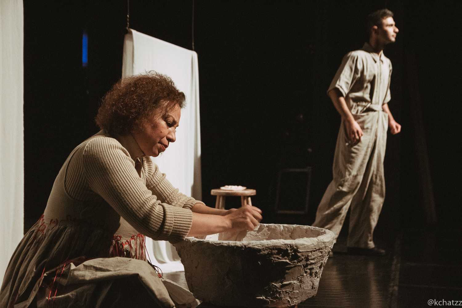 A woman kneels scrubbing her hands in a large stone basin as a man in overalls stands watching in the background