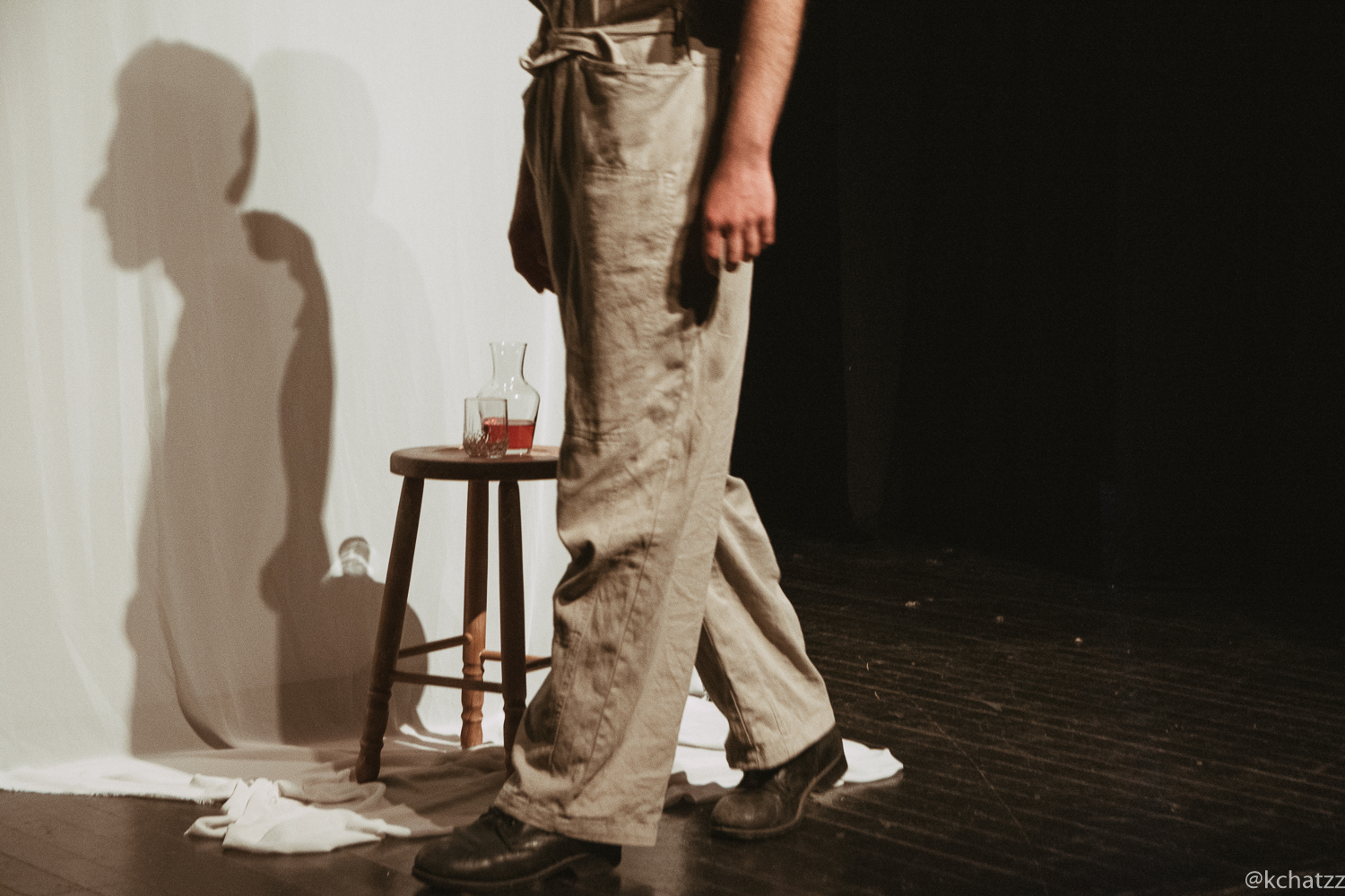 A performer's legs and boots pass a wooden stool holding a carafe and glass of red liquid, their shadow cast in profile on the curtain behind