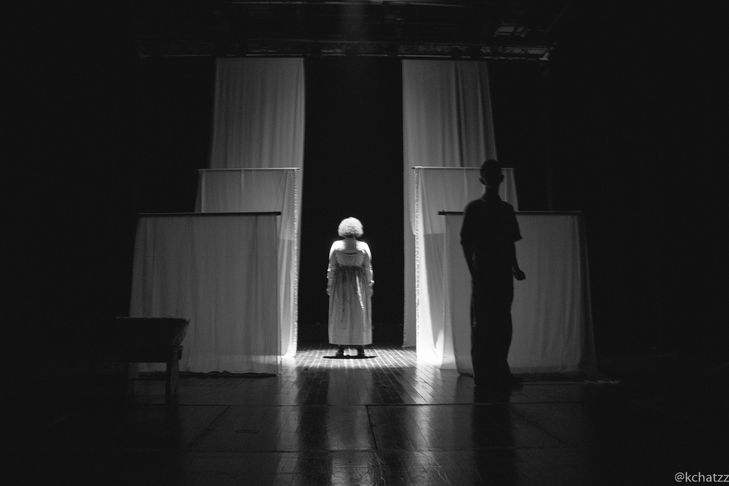 A woman in a white dress stands lit between hanging white curtain panels, with a man's silhouette watching from the side