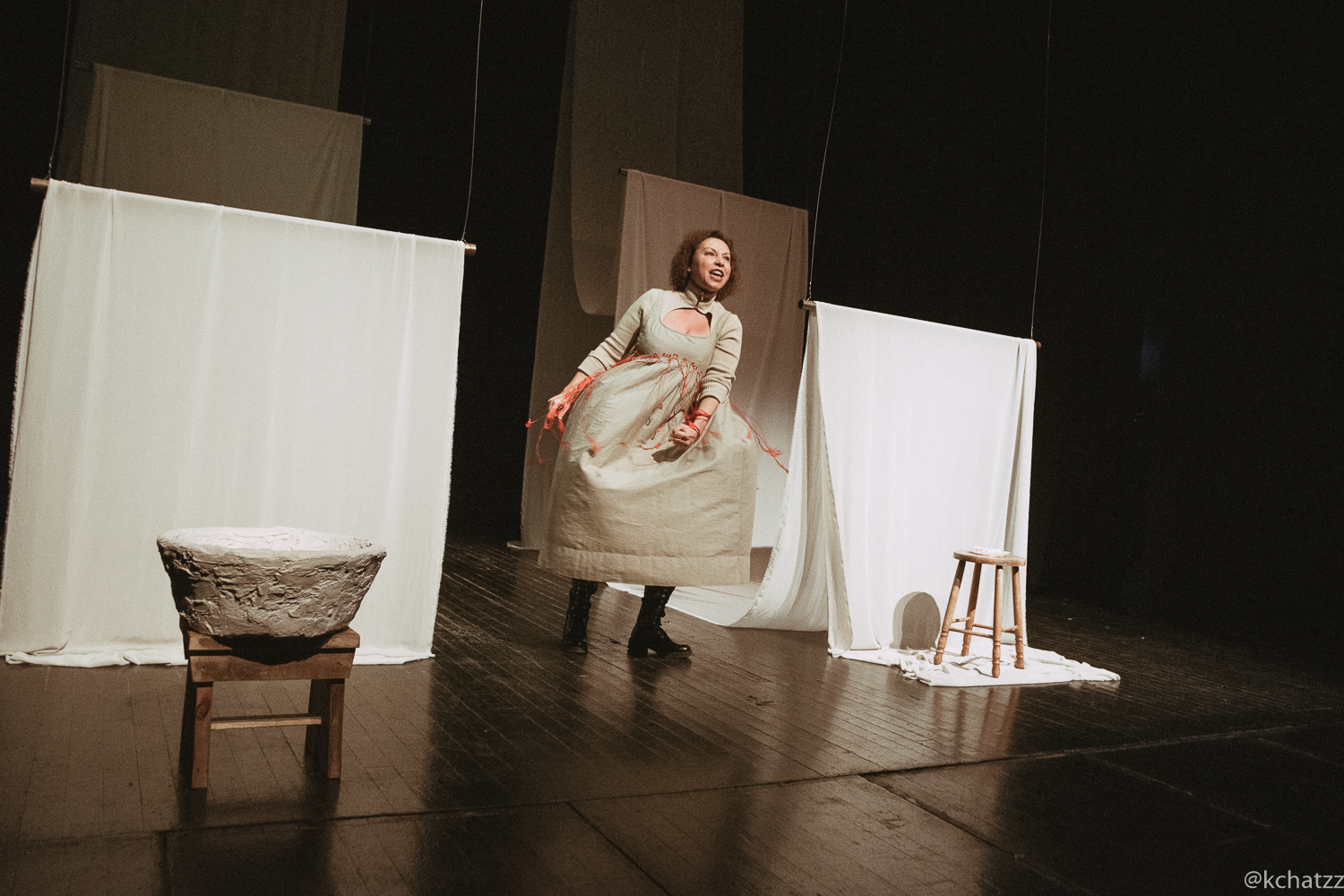 A woman in a beige dress trailed with red threads leans forward mid-speech between hanging curtains, beside a rough stone basin