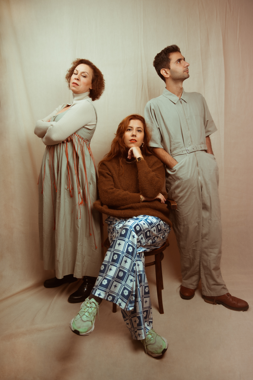 The three cast members pose together against a cream backdrop, one woman with arms crossed, a seated woman resting her chin on her hand, and a man looking upward