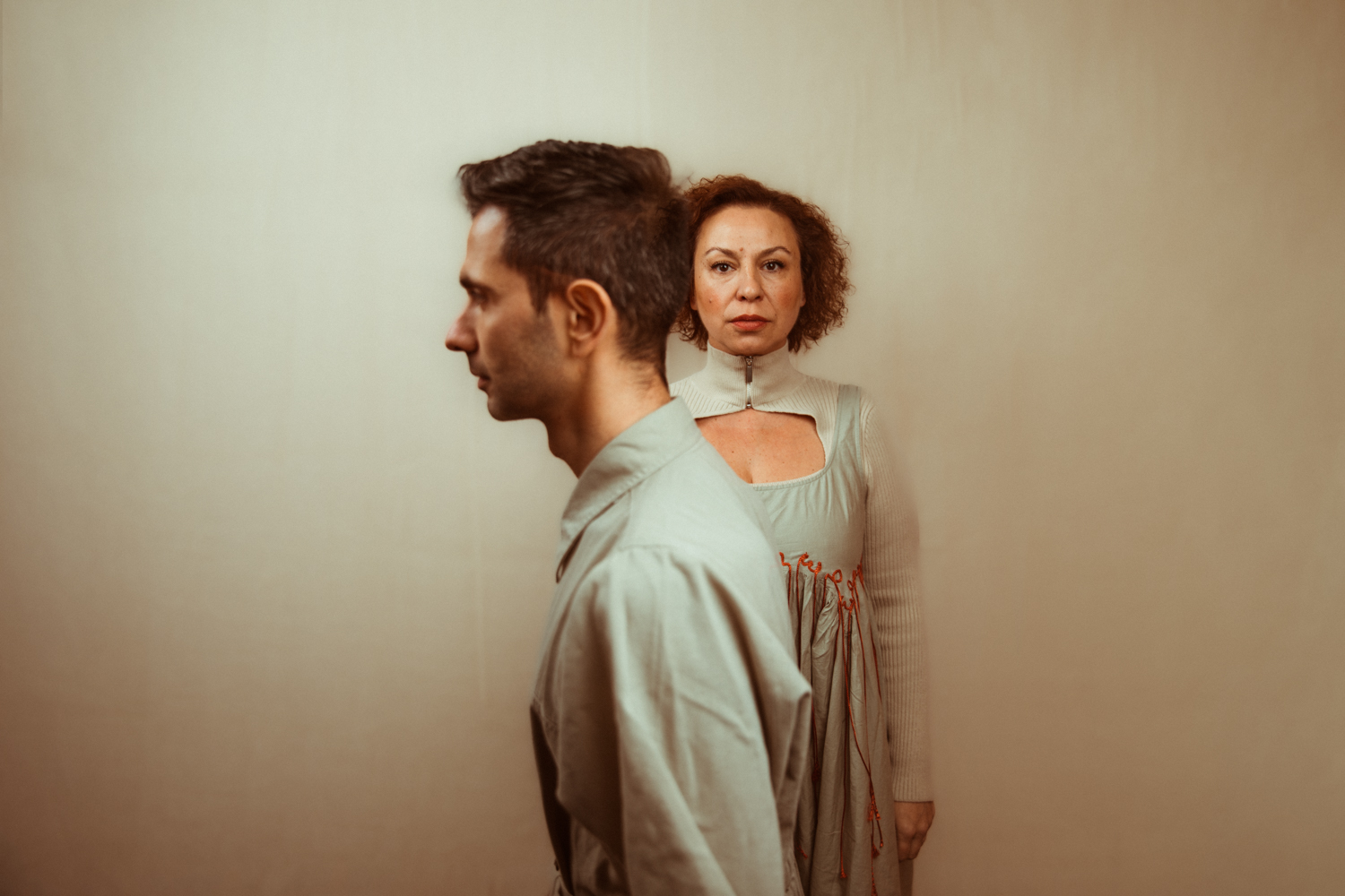 A man in profile stands back to back with a woman in a pale dress trimmed with red thread, both looking in opposite directions