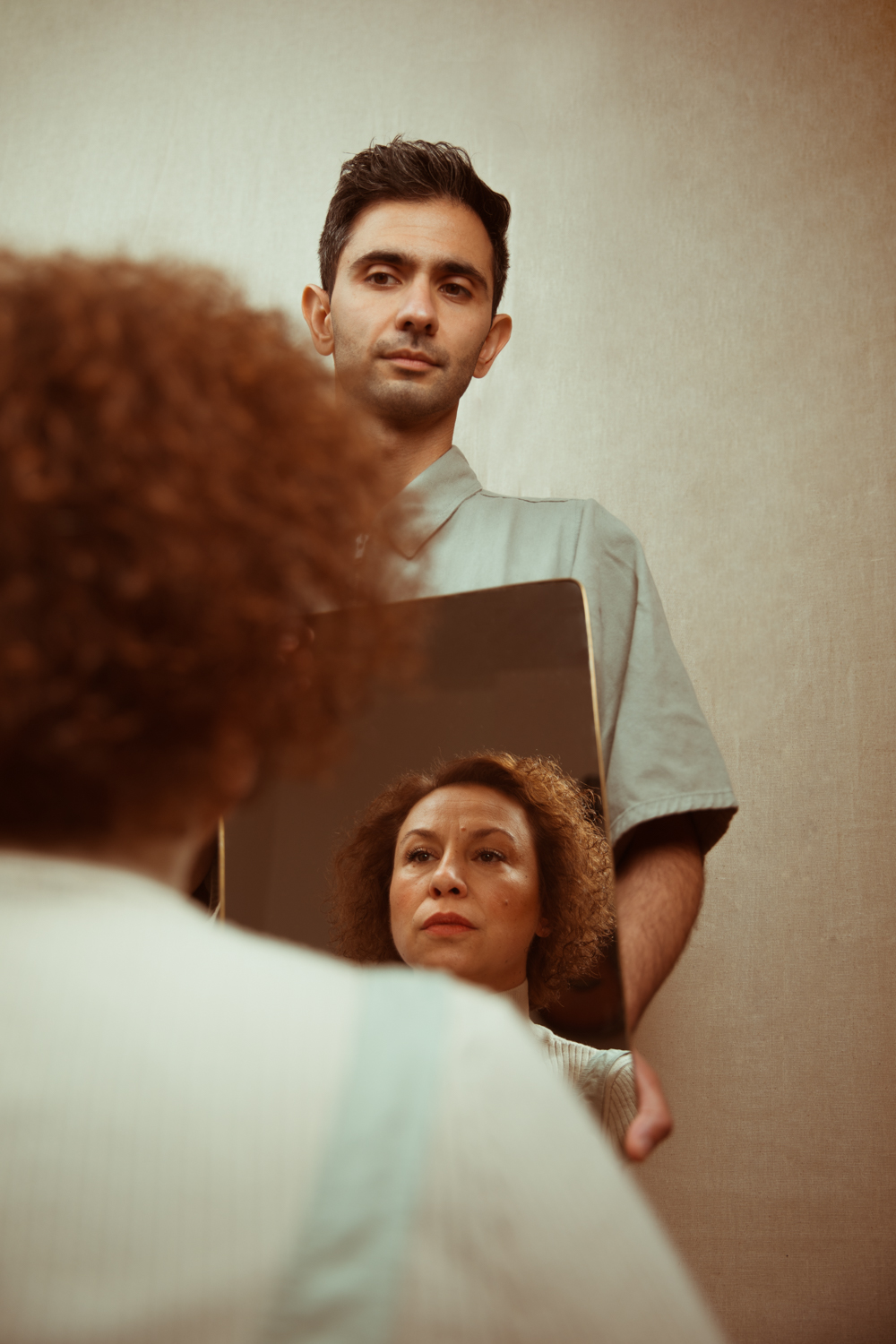 A man holds a small mirror reflecting a woman's face, her curly hair visible in the foreground