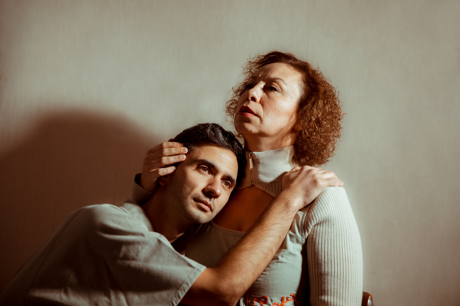 A man rests his head against a woman's shoulder as she holds his head, his hand resting on her shoulder in return