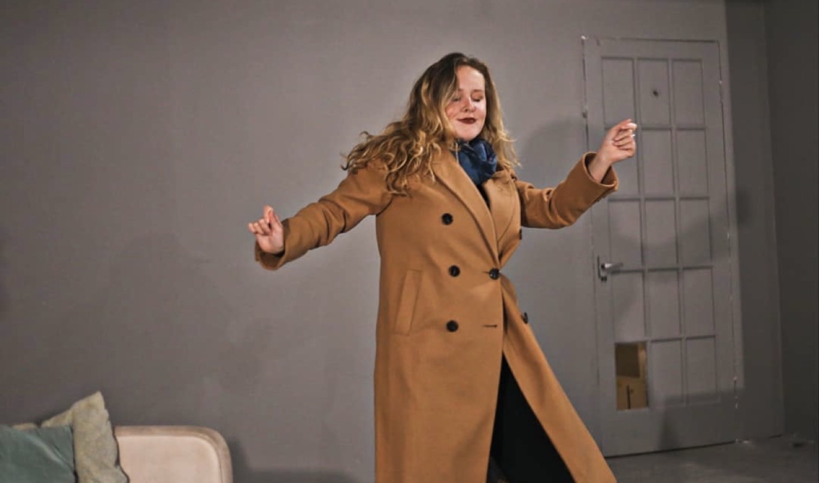 A performer in a camel coat dances with arms outstretched in front of a paneled door