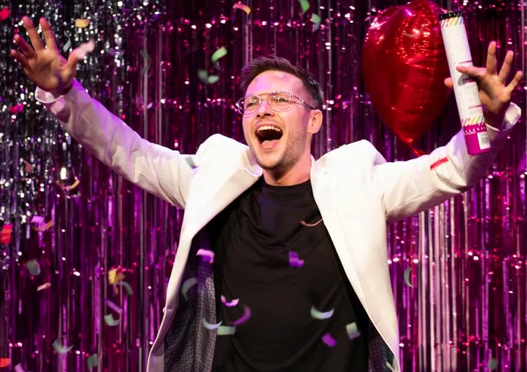 A performer throws his arms up celebrating amid confetti and a red balloon against a pink tinsel backdrop for First Time