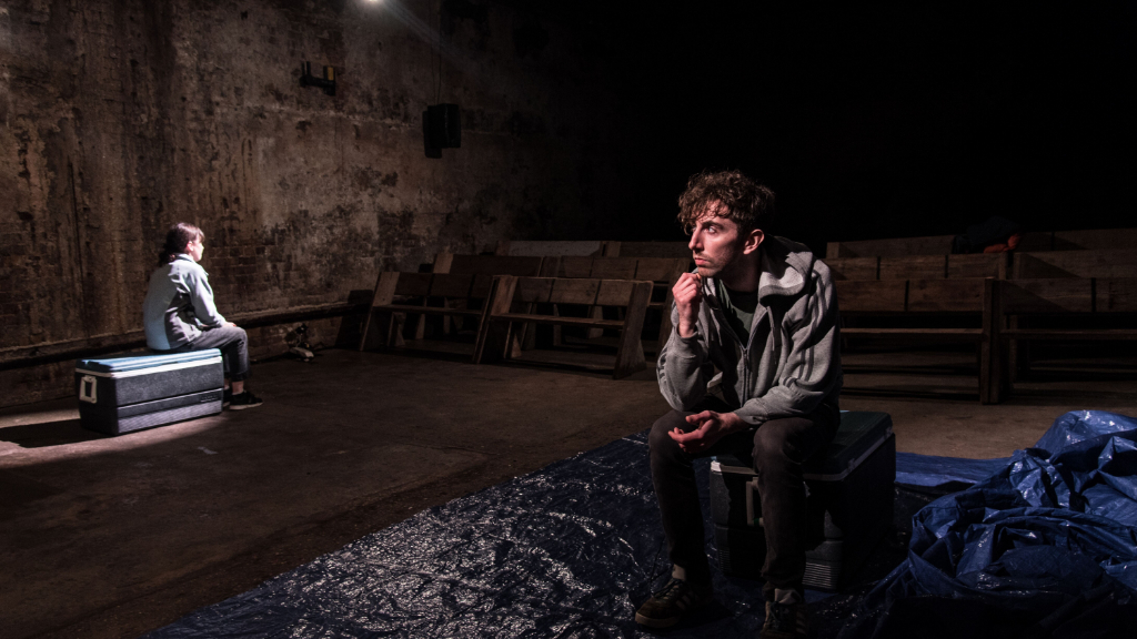 Two performers sit apart in a bare stone venue, one under a spotlight beside a blue tarpaulin, for In My Lungs The Ocean Swells