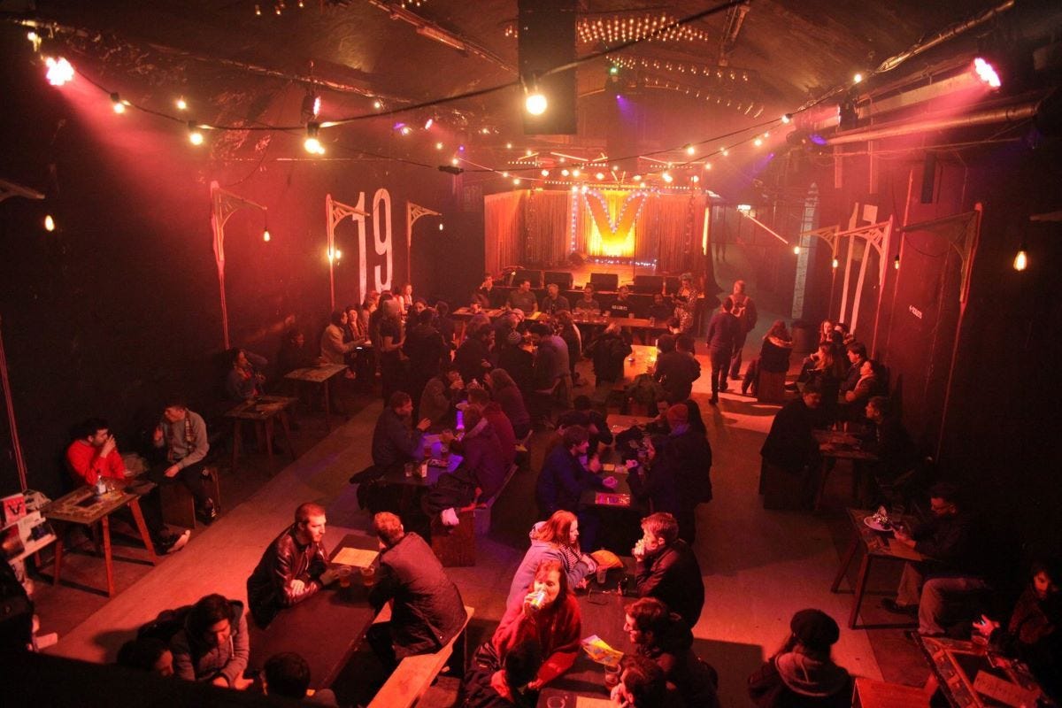 An overhead view of the Vaults venue tunnel, audience seated at tables under string lights facing a lit stage marked with a large V