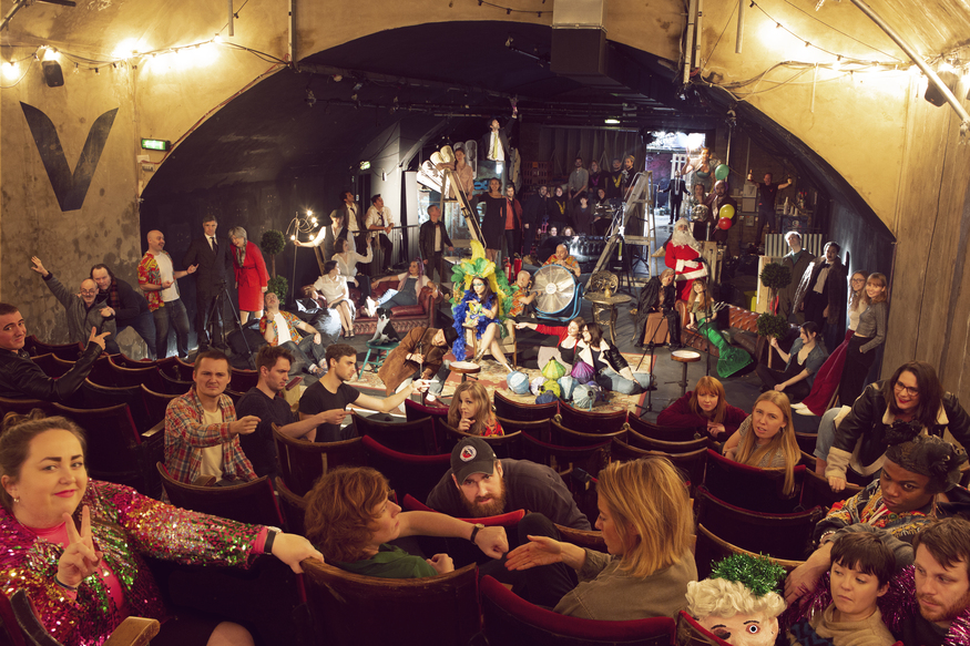 A large group of performers and press pose together in the tiered seating and stage area of the Vaults venue for the festival launch