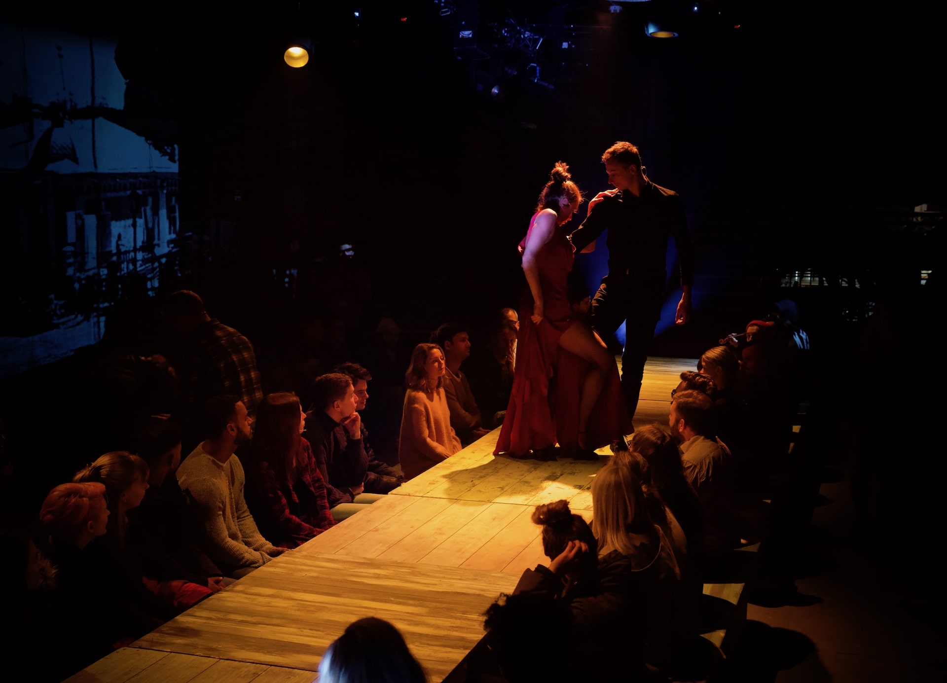 Two performers in red and black hold each other on a wooden promenade runway, flanked by seated audience members, with archive footage projected on the wall behind