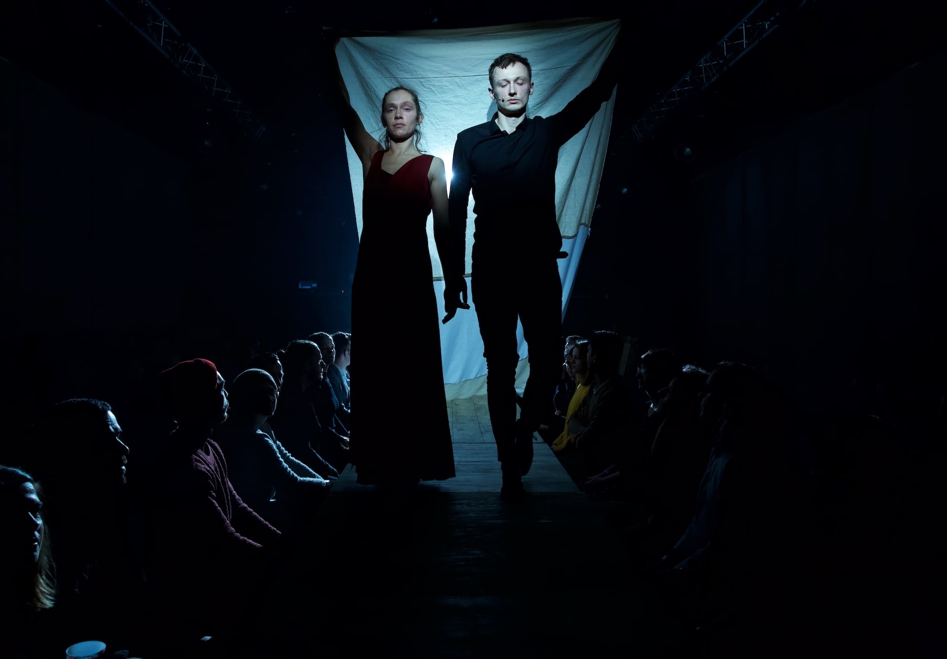 A performer in a red dress and a performer in black stand together holding up a backlit white sheet above a promenade audience