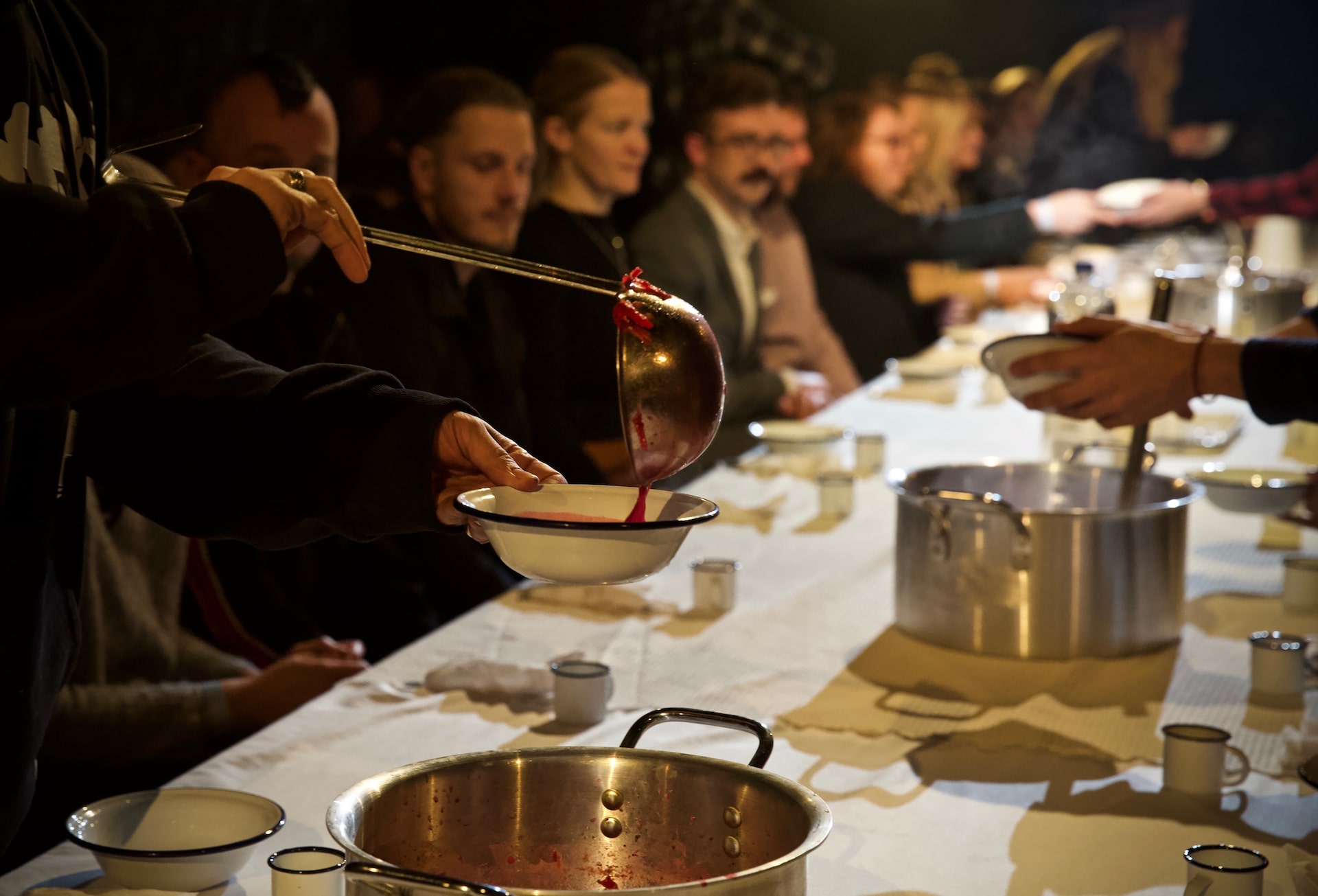 A hand ladles red liquid into an enamel bowl held by an audience member seated at a long communal table set with pots and cups