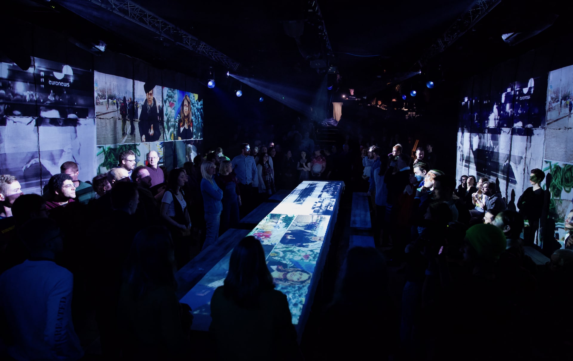 A wide view of the promenade space, audience standing on either side of a long illustrated runway beneath archive news footage projected across both walls