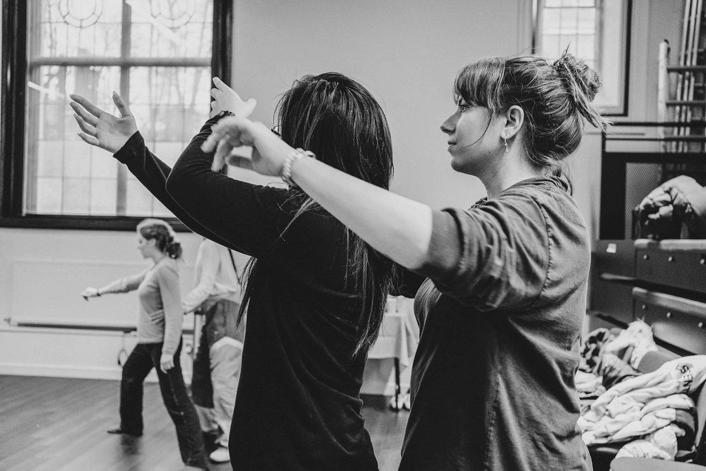 Two performers rehearse a partnered movement in a studio, arms raised in unison, as others move in the background