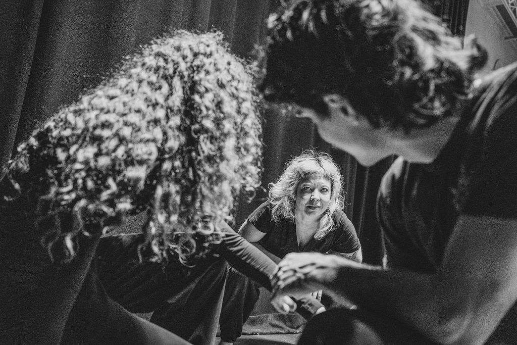 Three performers share a close moment on stage, one seated and looking up as two others lean in with joined hands