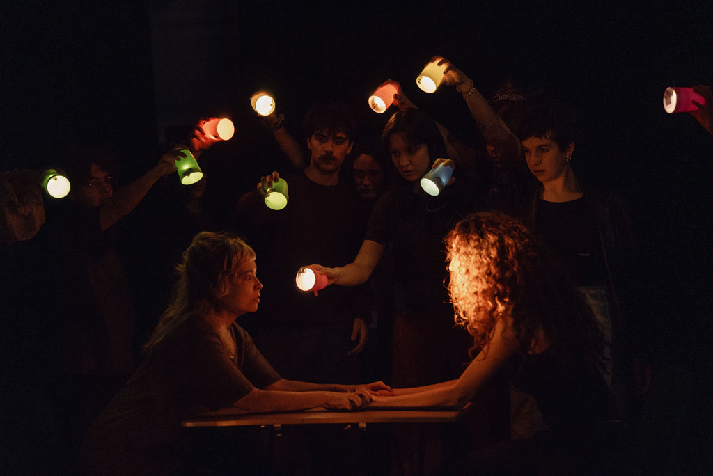 Two performers sit face to face at a table lit only by glowing paper cups held up by a circle of performers around them