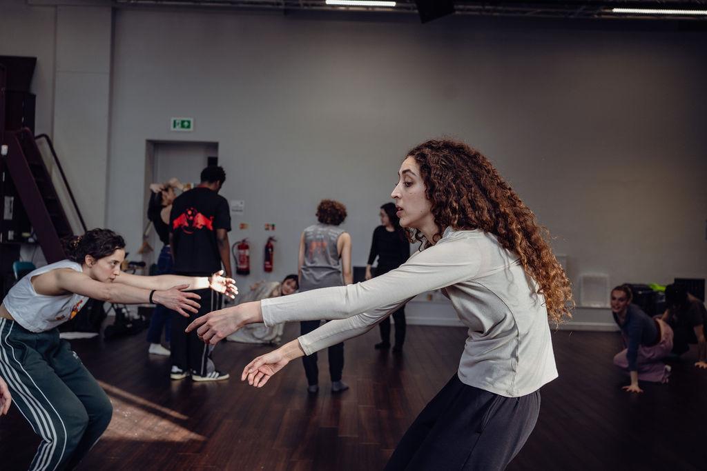 A performer with long curly hair reaches both arms forward mid-movement as other dancers move around her in a bright studio