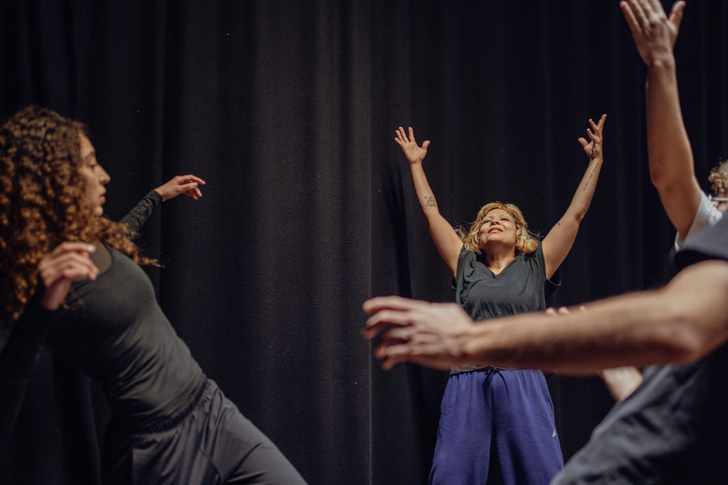 A performer raises both arms overhead with her face turned up to the light, flanked by other performers reaching outward on a dark stage