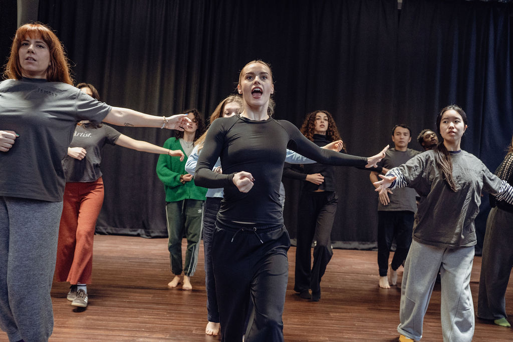 A group of performers advance across the studio floor with arms outstretched, one woman in front calling out mid-stride