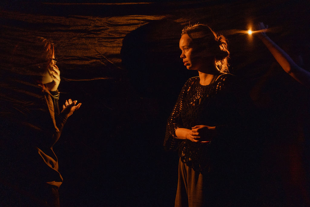 Two performers face each other in warm firelight against a dark draped backdrop, one gesturing with an open hand