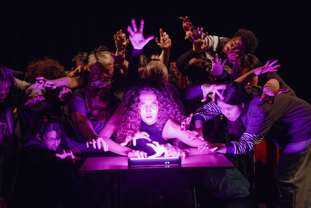 A performer lit in purple light reaches toward a glowing lamp on a table, surrounded by a crowd of performers with hands raised