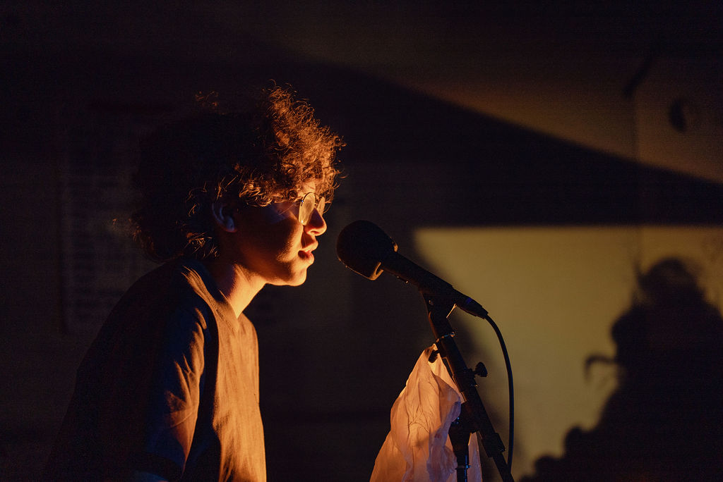 A performer lit in warm light sings into a microphone stand draped with white fabric against a dark backdrop
